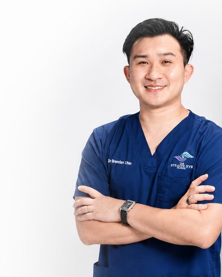 Dr Brendan Chin in navy scrubs with his arms folded, facing the camera against a plain studio backdrop.