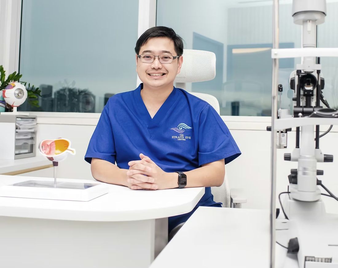 Dr Jason Lee at a consultation desk in Straits Eye Centre scrubs, with a slit lamp beside him.