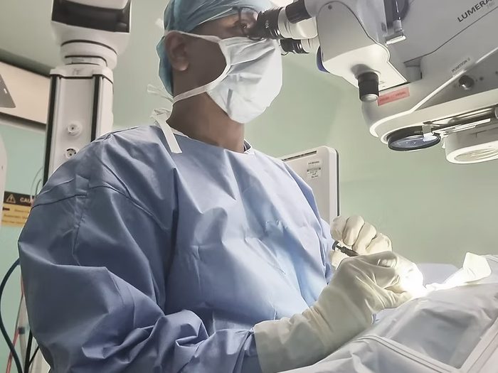 Dr Jayant V Iyer, gowned and masked, operating at a surgical microscope with an instrument in his gloved hand.
