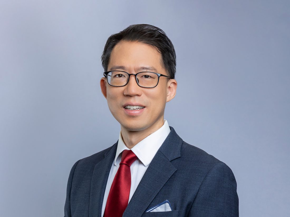 Dr Laurence Lim in a navy suit and red tie, photographed against a plain grey backdrop.