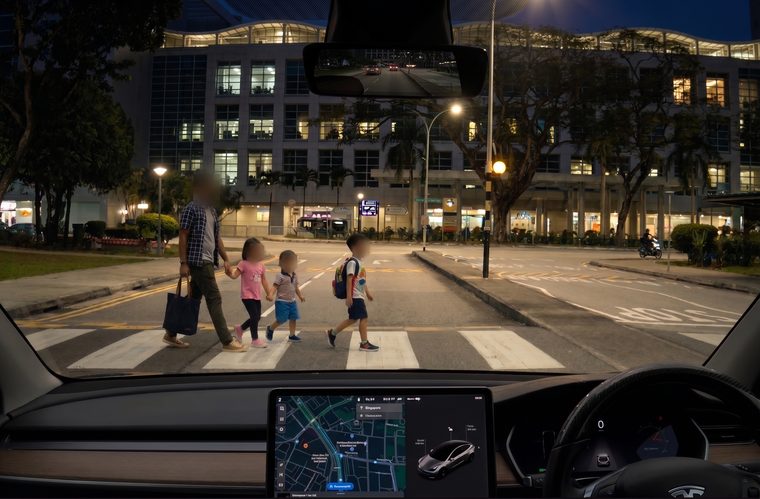 A driver&rsquo;s view through a windscreen at night: a man and three children crossing at a zebra crossing ahead, in sharp focus under street lighting.
