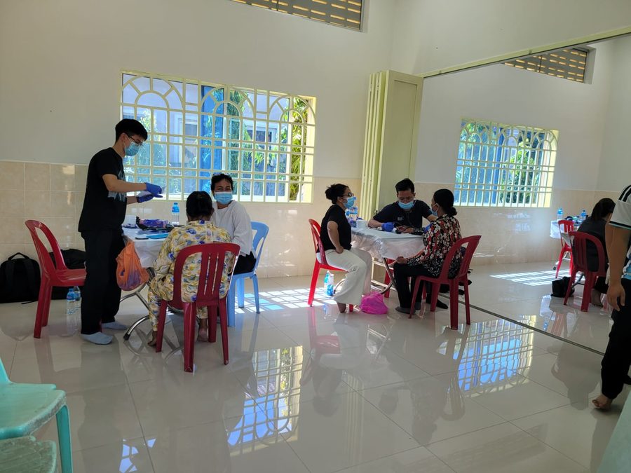 Masked clinic staff seeing seated patients at folding tables in a tiled hall.