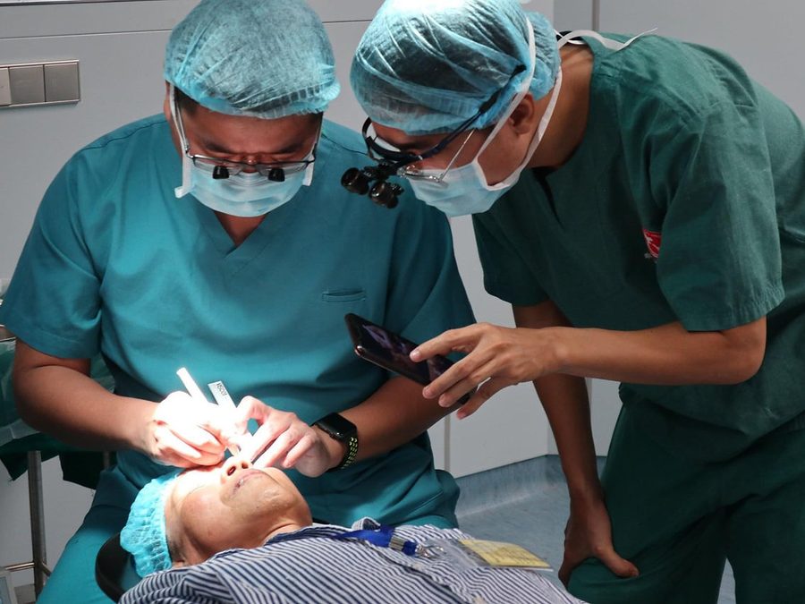 Two surgeons in scrubs, caps and surgical loupes operating on a patient&rsquo;s eye, with a colleague recording on a phone.