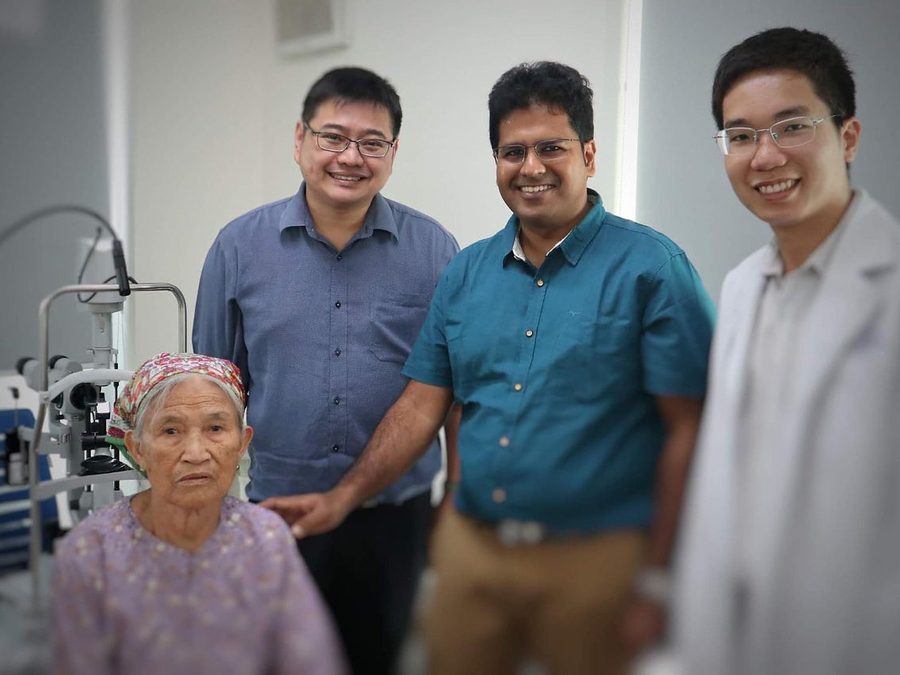 Dr Jason Lee and Dr Jayant V Iyer with a colleague in a white coat and a seated patient, beside a slit lamp in an eye clinic in Hanoi.